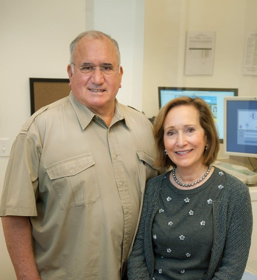 A man in a beige short-sleeve shirt stands next to a smiling woman in a gray sweater and pearl necklace; they are indoors, with computer monitors and office equipment in the background.