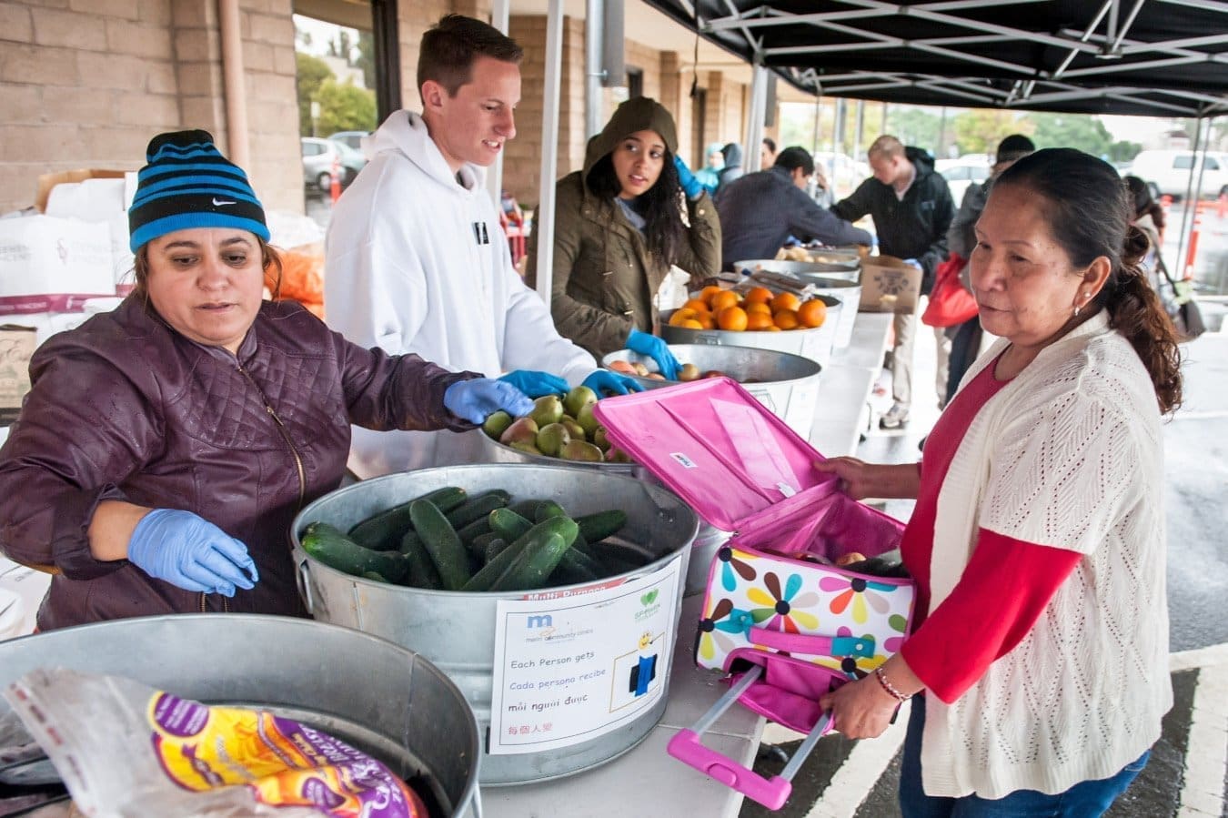 Several people stand at an outdoor market table handing out fresh vegetables and fruits to a woman with a pink shopping bag. Some wear gloves and jackets, and the setting appears to be a community food distribution event.