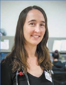 A woman with long brown hair wearing a black top and a stethoscope around her neck smiles in an office or medical setting, with blurred people and equipment in the background.