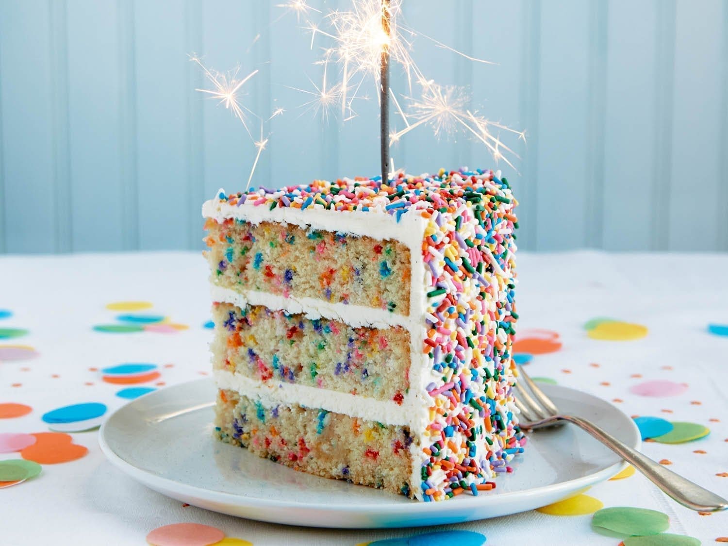 A slice of three-layer funfetti cake with white frosting and colorful sprinkles, topped with sparklers, sits on a white plate with a fork. The table and background are decorated with colorful confetti.