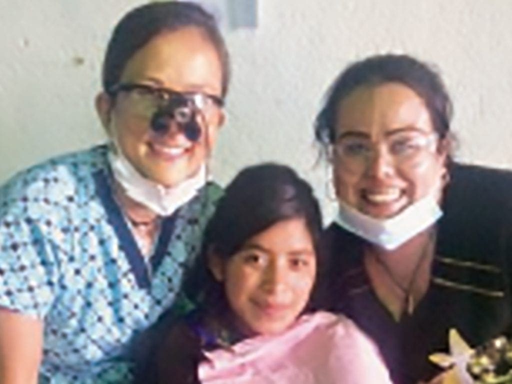 Two women in medical attire, both wearing glasses and masks under their chins, smile while standing beside a young girl with long dark hair, wrapped in a pink blanket. All three appear happy and are indoors.