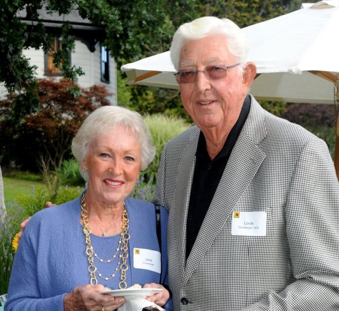 An older woman and man stand together outdoors, both smiling at the camera. The woman holds a teacup and wears a blue jacket and gold necklace. Both wear name tags and the background shows greenery and a white umbrella.