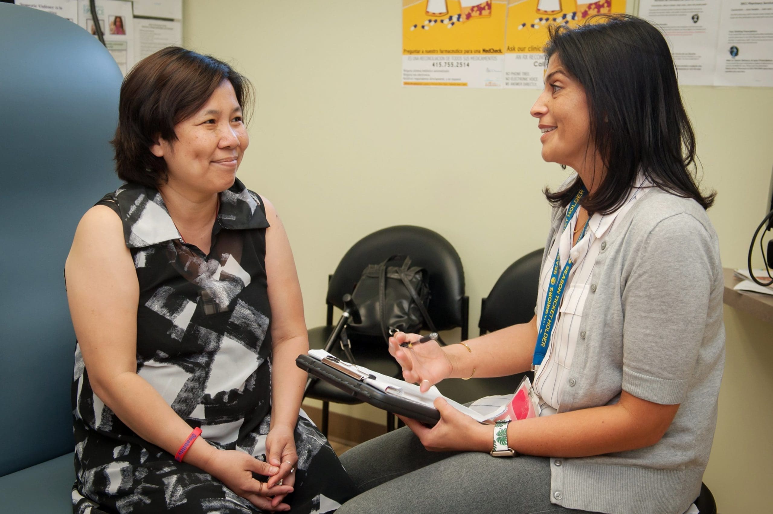 A female doctor sits and speaks with a smiling female patient in a medical office. The doctor holds a clipboard and pen, engaging in conversation. Medical posters and chairs are visible in the background.
