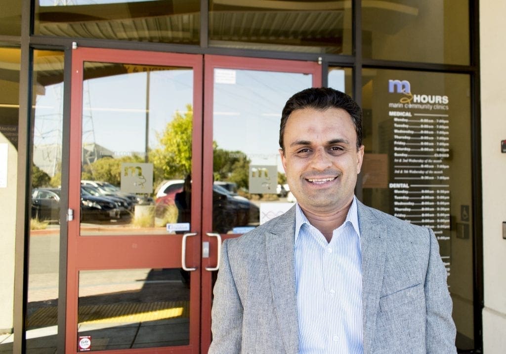 A man in a light gray suit jacket and striped shirt stands smiling in front of glass doors at a clinic entrance, with clinic hours posted on the window behind him.