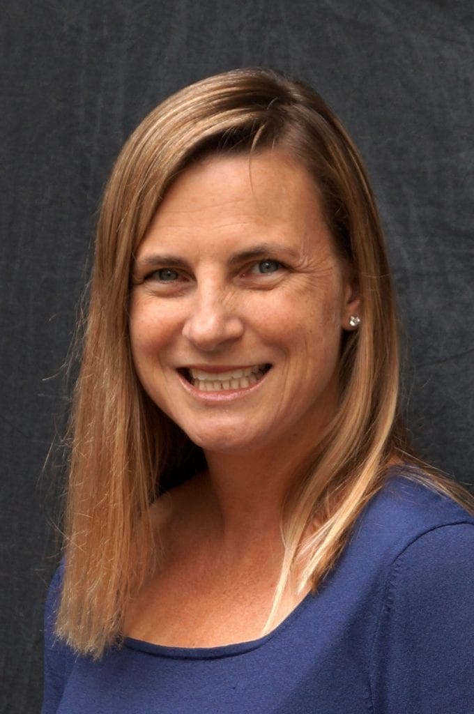 A woman with straight, shoulder-length blonde hair and blue eyes smiles at the camera. She is wearing a blue top and small stud earrings, posing in front of a dark, plain background.
