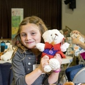 A smiling young girl holding a white teddy bear dressed in red clothes, sitting indoors at a table with other people and craft materials in the background.