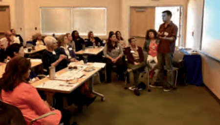 A man stands and speaks to a group of seated adults in a classroom or meeting room, with a whiteboard or projection screen behind him. The group appears attentive and engaged.