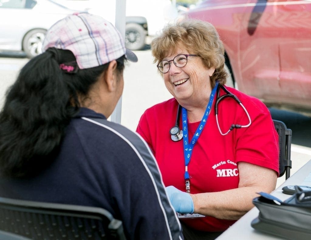 A smiling healthcare worker in a red shirt, wearing a stethoscope, talks to a woman in a plaid cap at an outdoor table. Medical equipment is on the table, and cars are visible in the background.
