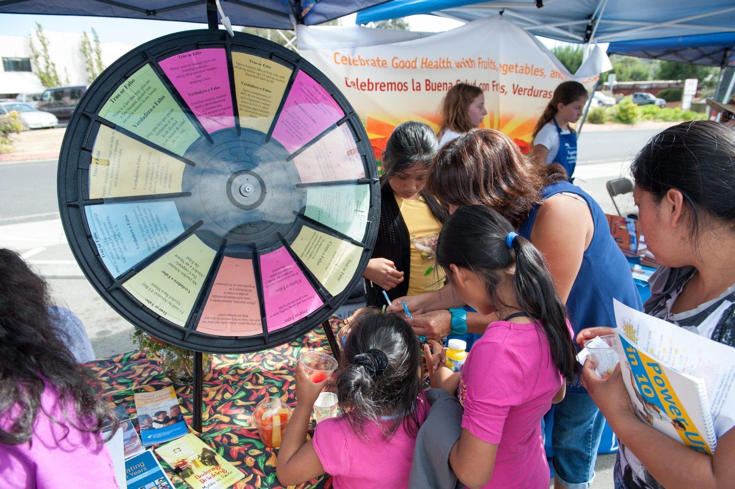 A group of people, including children and adults, gather around a colorful prize wheel at an outdoor health fair booth, with informational banners and handouts visible.
