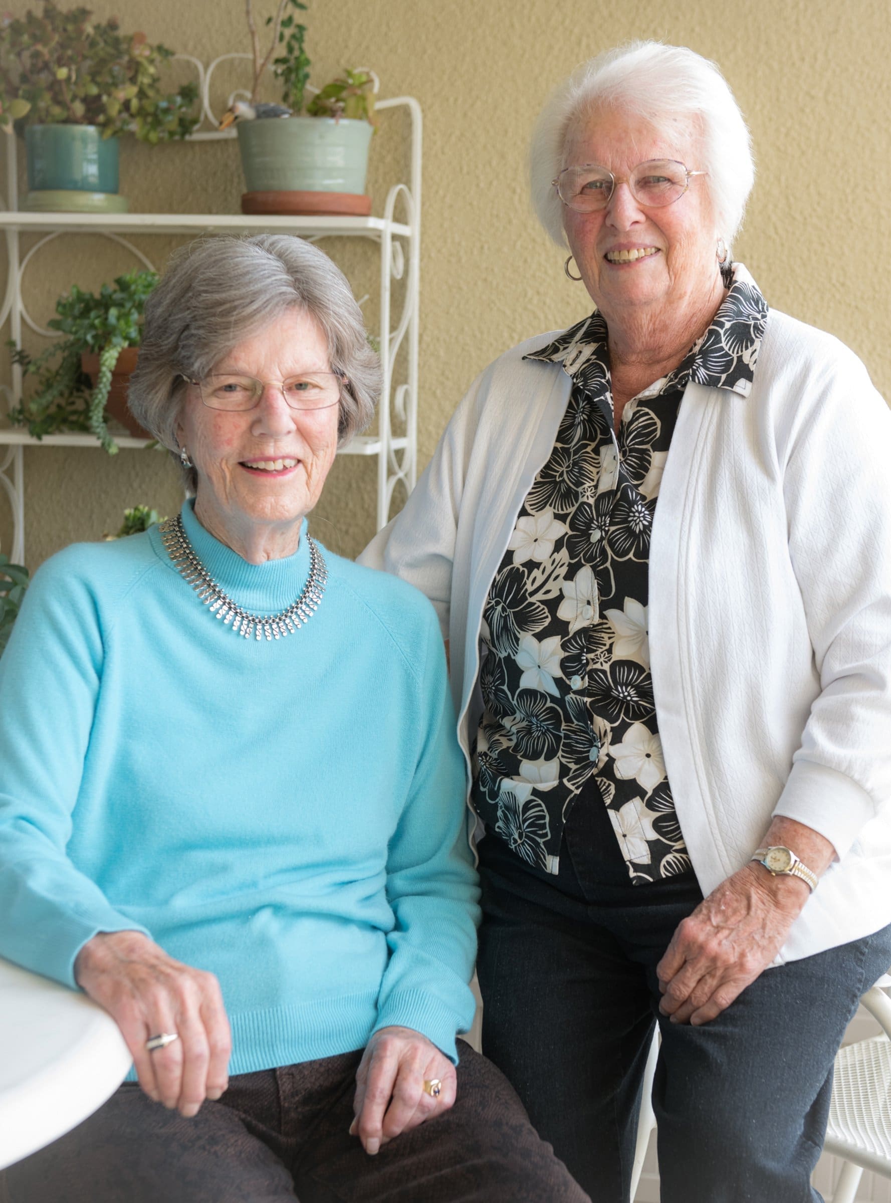 Two elderly women smile together indoors; one is seated in a blue sweater, the other stands beside her in a white cardigan and floral shirt. Potted plants are in the background.