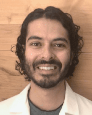 A person with medium-length curly hair and a beard smiles while wearing a white lab coat, standing in front of a wooden wall.