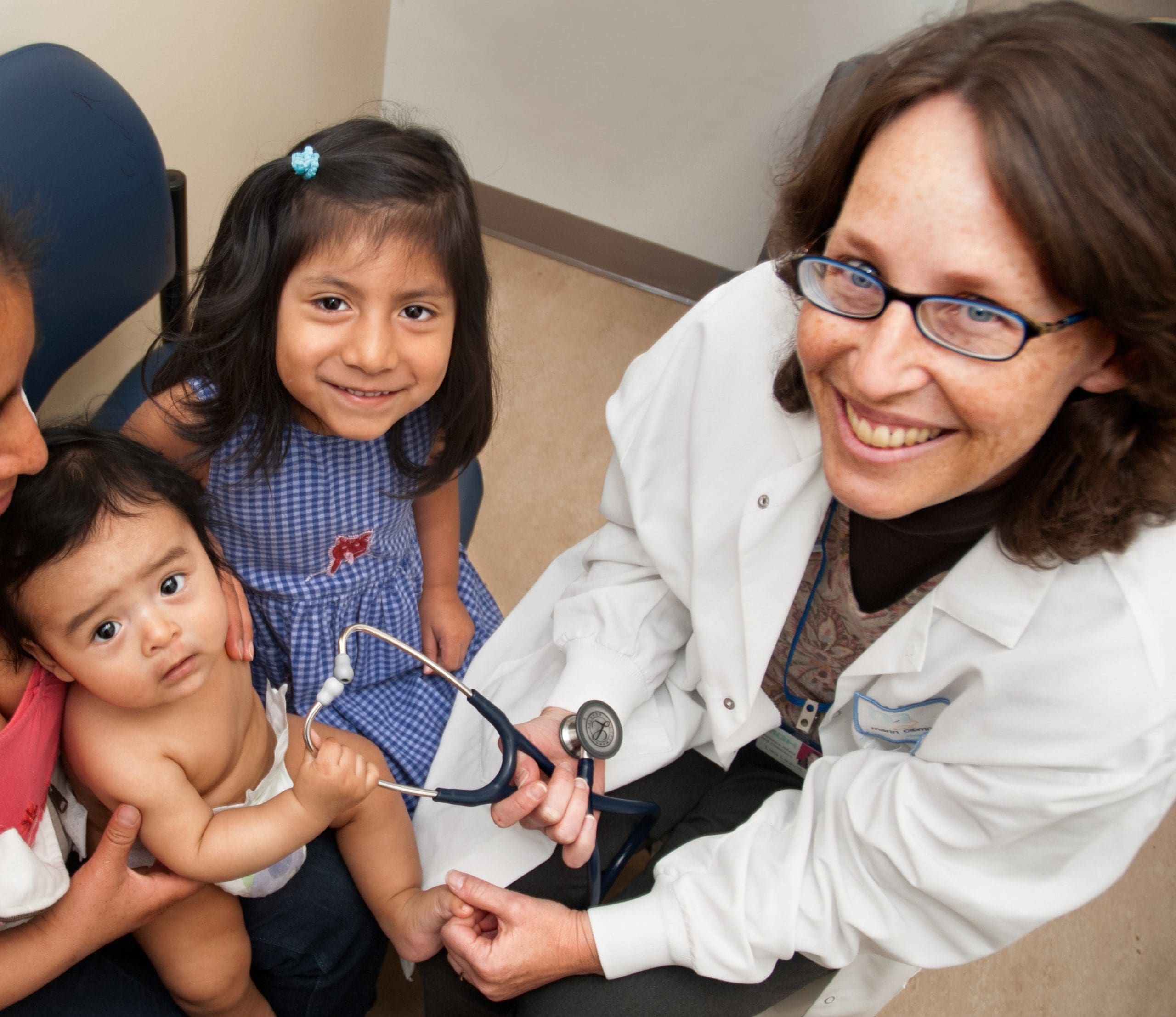 A smiling doctor in a white coat holds a stethoscope next to a baby and a young girl, who are both sitting on an adults lap. The young girl is smiling at the camera. The scene takes place in a medical office.