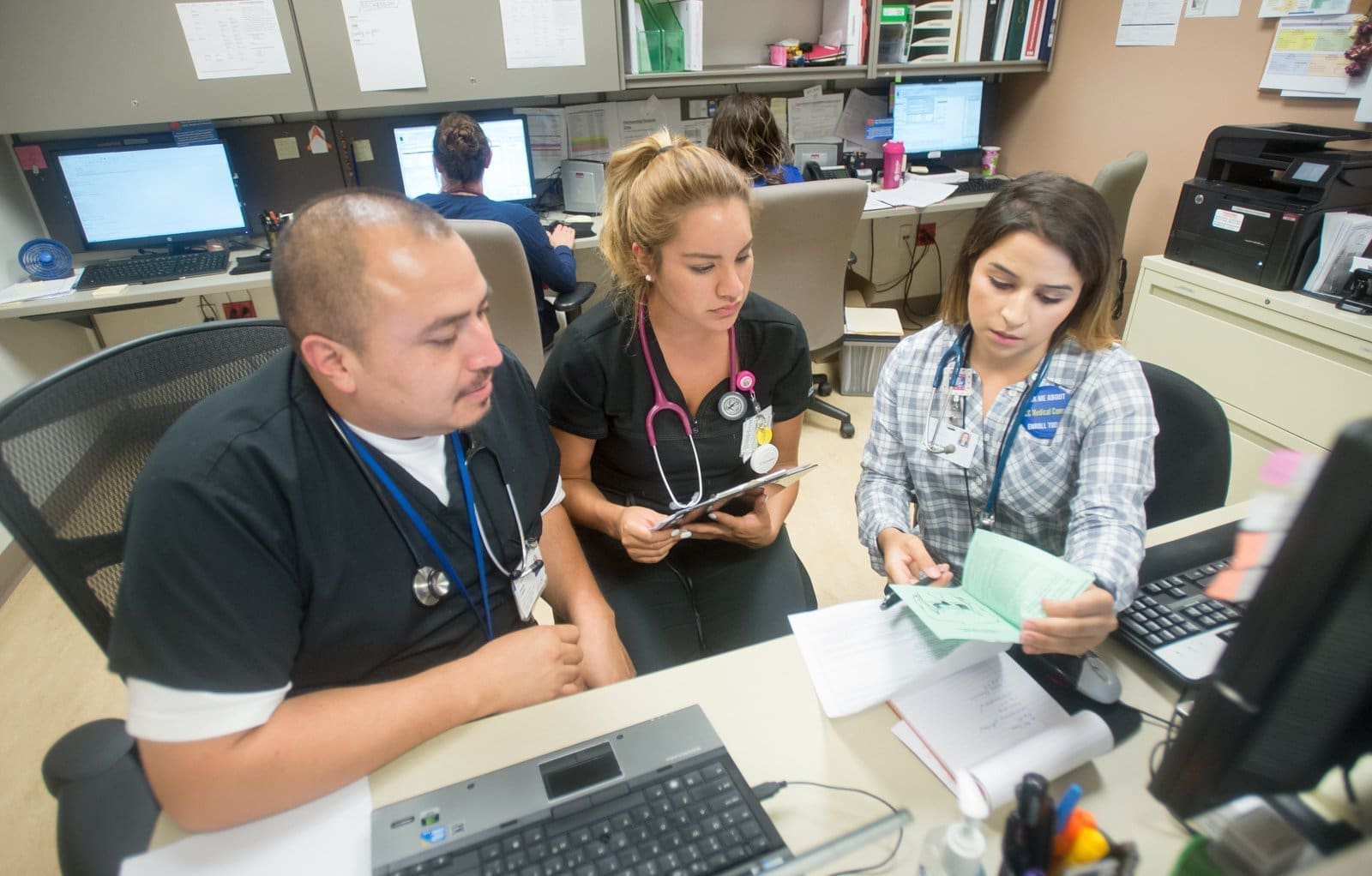 Three healthcare professionals sit at a desk in an office, reviewing documents together. Computer monitors, paperwork, and office supplies are visible around them, and two more people work at computers in the background.