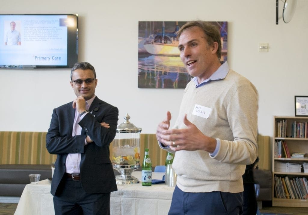 Two men are standing indoors near a table with drinks. One man is speaking while gesturing with his hands, and the other stands with his hand on his chin, listening. A painting and TV screen are visible in the background.