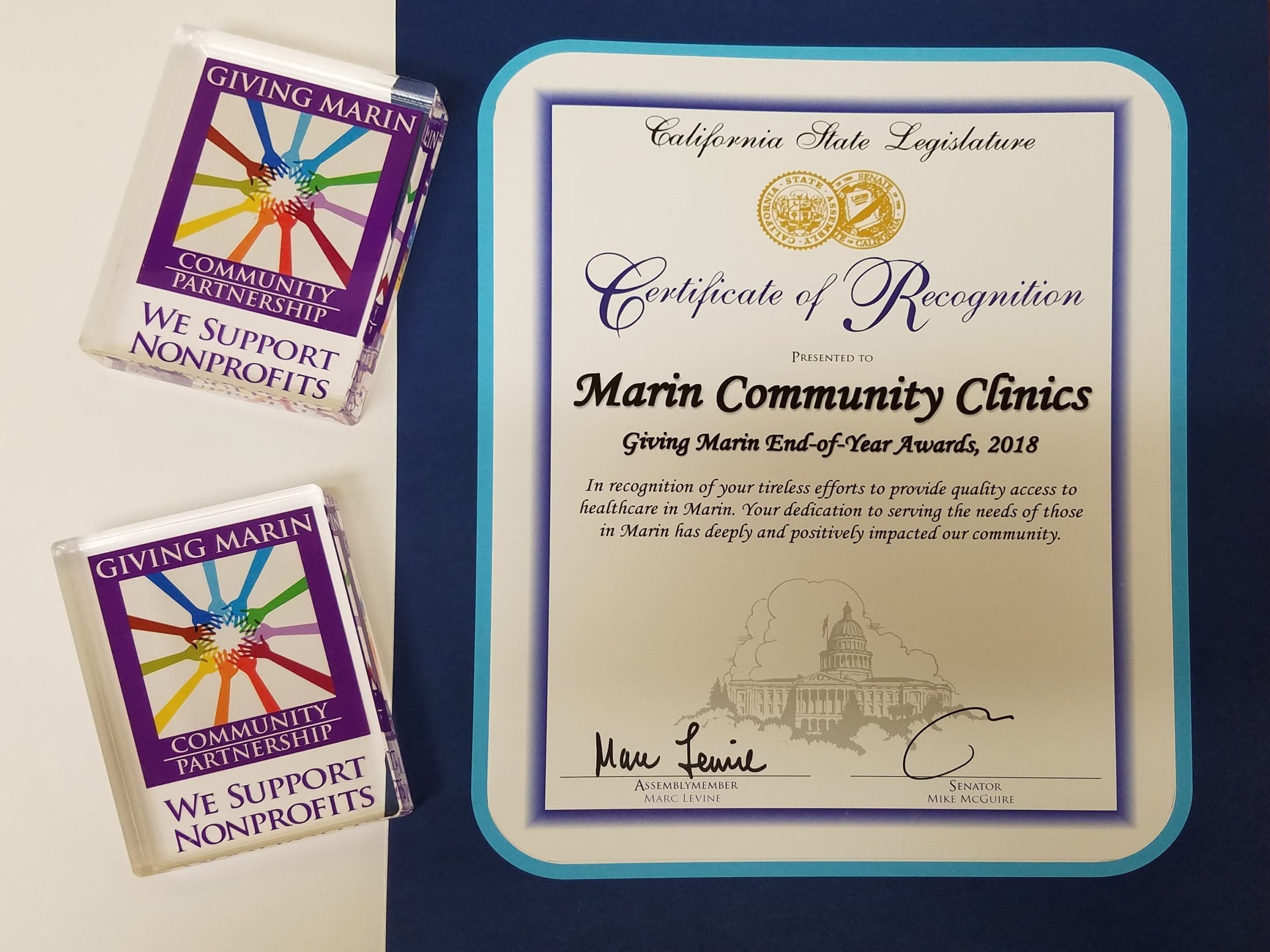 A Certificate of Recognition from the California State Legislature awarded to Marin Community Clinics for the 2018 Giving Marin End-of-Year Awards, alongside two colorful We Support Nonprofits badge holders.