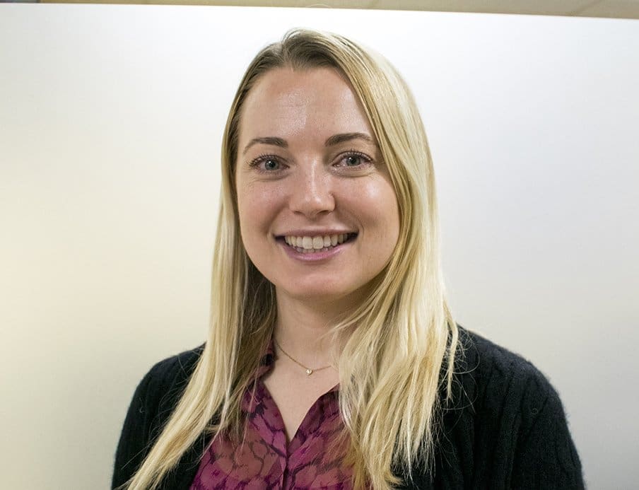 A woman with long blonde hair, wearing a dark sweater over a purple patterned blouse, smiles at the camera against a plain, light-colored background.