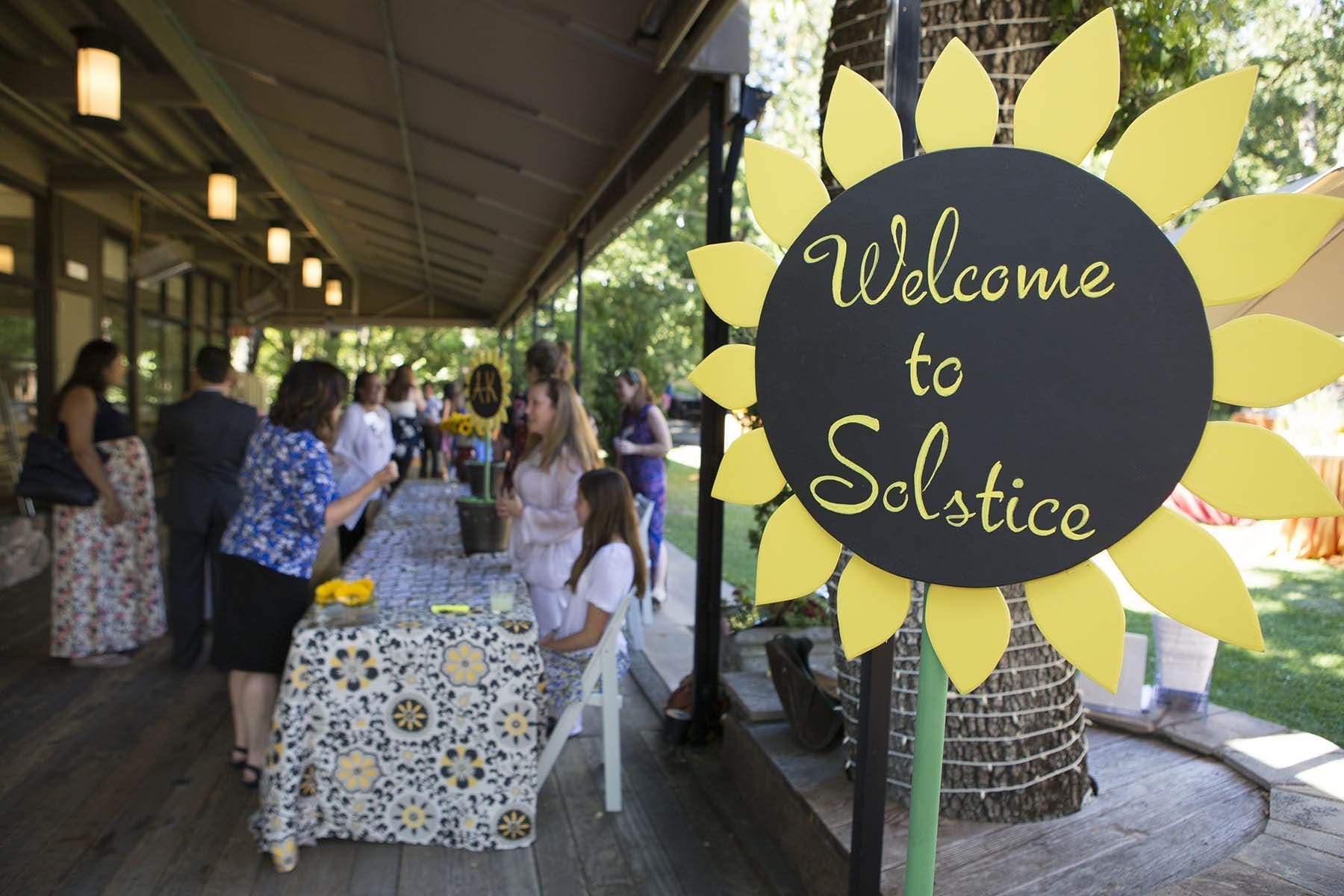 A large sunflower-shaped sign reading Welcome to Solstice stands near a covered outdoor area where people are gathered around tables decorated with floral-patterned cloths. Trees and greenery are visible in the background.