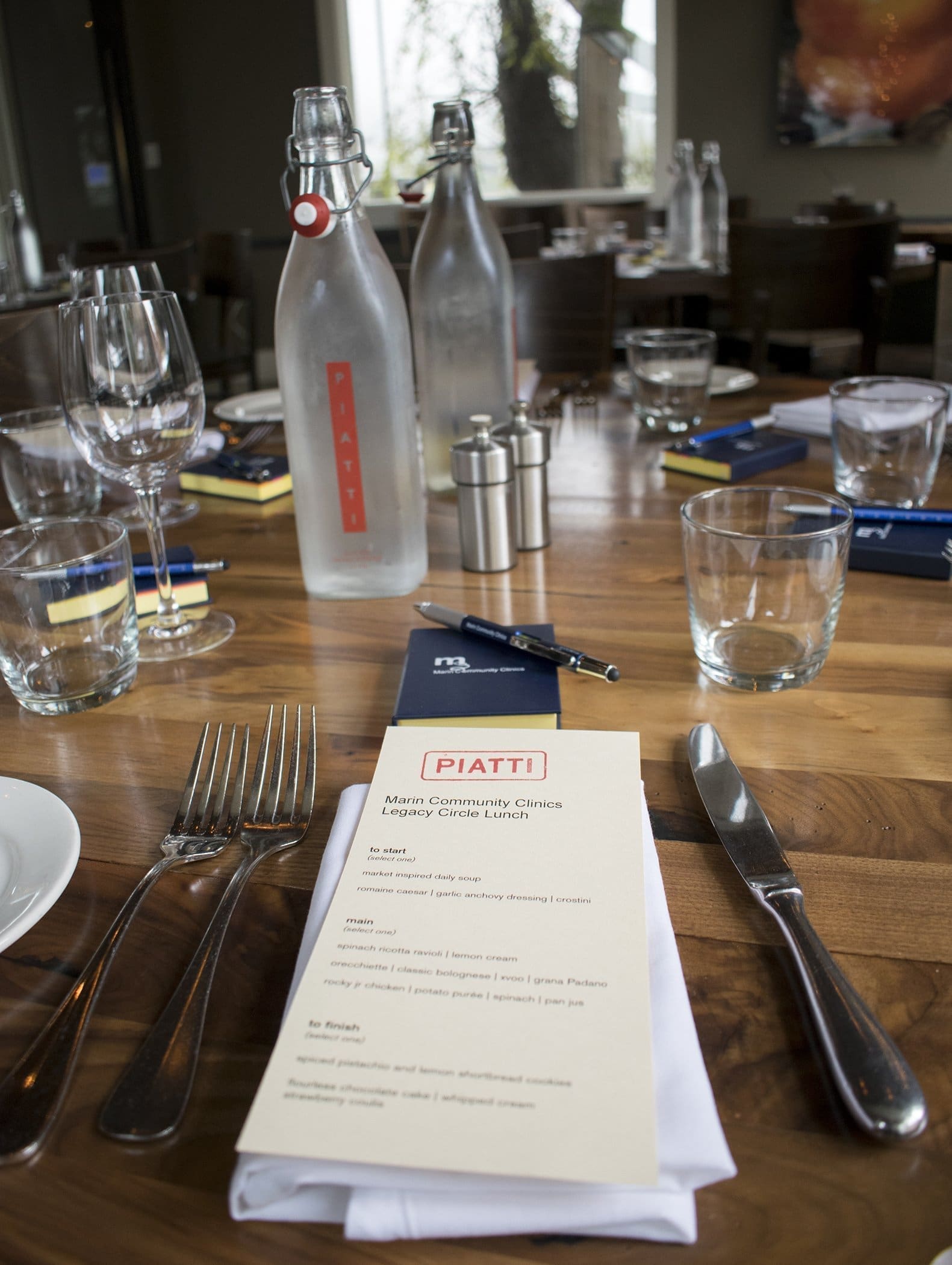 A wooden restaurant table set with glasses, cutlery, napkins, two glass bottles of water, and a menu labeled Piatti. The background shows more tables, chairs, and large windows.