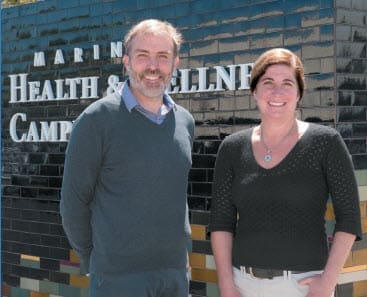 Two people stand smiling in front of a black tiled wall with the words Marin Health & Wellness Campus partially visible in white letters.