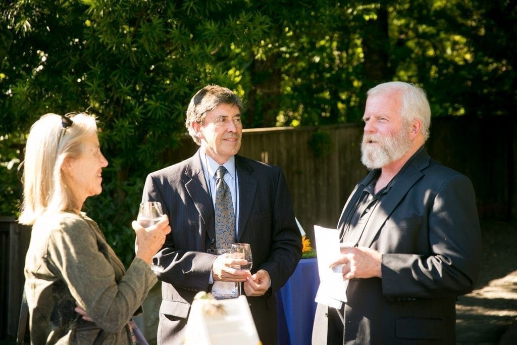 Three people wearing suits and holding drinks stand outdoors, engaged in conversation about the recent news of a cancer diagnosis, with sunlight filtering through green trees in the background.