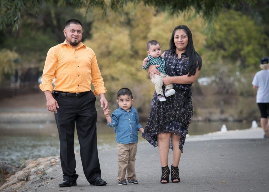 A family of four stands on a park path by a pond, sharing a quiet moment after the news of a cancer diagnosis. The father, in an orange shirt, holds his young son's hand while the mother, in a dark dress, carries their baby. Trees and water fill the background.