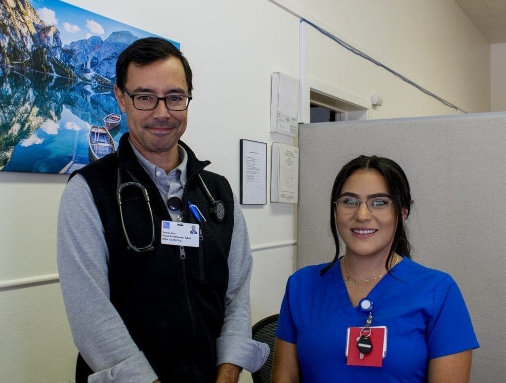 A man in a black vest and stethoscope stands next to a woman in blue scrubs; both are smiling in an office with a scenic mountain lake photo on the wall, reflecting their commitment to integrated care.
