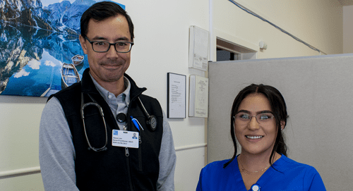 A male doctor with glasses and a stethoscope stands beside a smiling female nurse in blue scrubs in a medical office. A mountain photo hangs on the wall behind them.