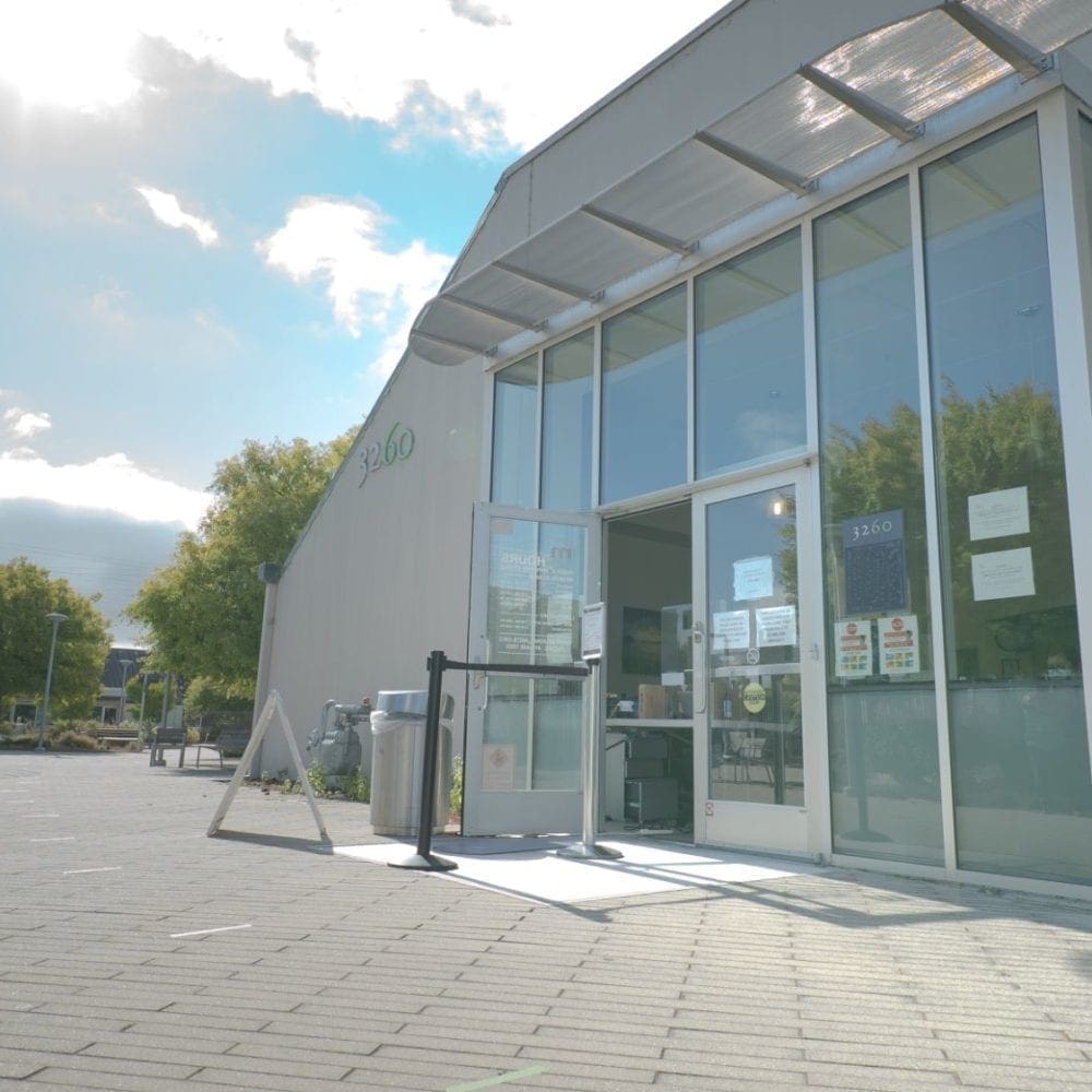 A modern building with large glass doors and windows, featuring signs and a hand sanitizer station at the entrance, is seen on a sunny day with a few clouds in the sky and some trees nearby.