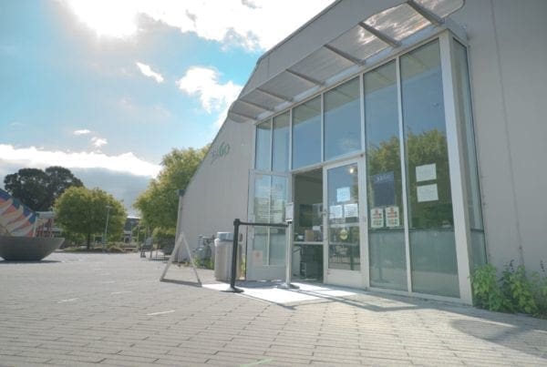 A modern building with large glass doors and windows, featuring signs and a hand sanitizer station at the entrance, is seen on a sunny day with a few clouds in the sky and some trees nearby.