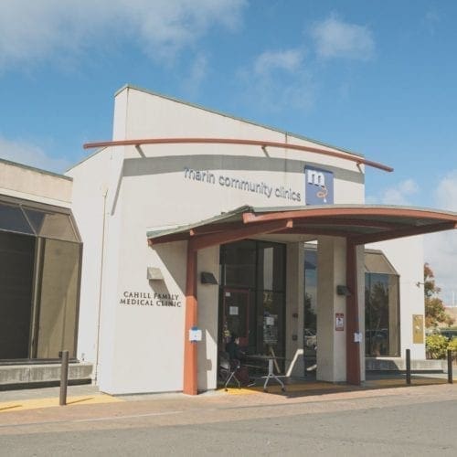 The image shows the entrance of Marin Community Clinics Carell Family Medical Clinic, a modern white building with large windows, a curved red awning, and a blue sky with clouds in the background.