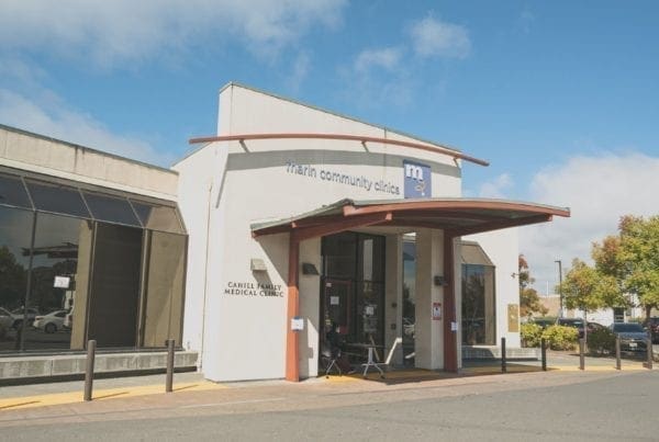 The image shows the entrance of Marin Community Clinics Carell Family Medical Clinic, a modern white building with large windows, a curved red awning, and a blue sky with clouds in the background.