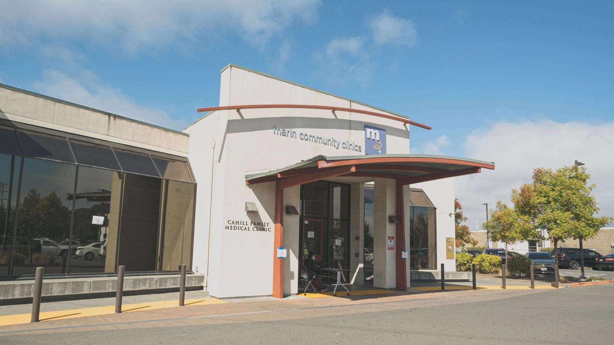 The image shows the entrance of Marin Community Clinics Carell Family Medical Clinic, a modern white building with large windows, a curved red awning, and a blue sky with clouds in the background.