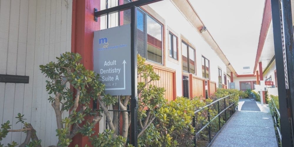 A walkway with a ramp leading to a building entrance. A sign reads Adult Dentistry Suite A with an arrow pointing forward. Bushes line the walkway, and the building is white with red trim and many windows.