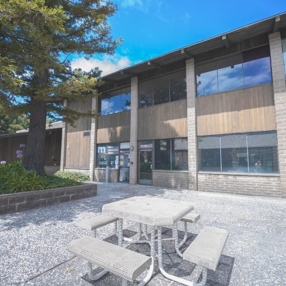 A concrete picnic table with benches sits in front of a two-story office building with large windows, brick pillars, and shaded by tall trees on a sunny day.