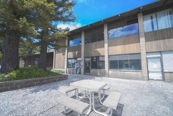 A concrete picnic table with benches sits in front of a two-story office building with large windows, brick pillars, and shaded by tall trees on a sunny day.