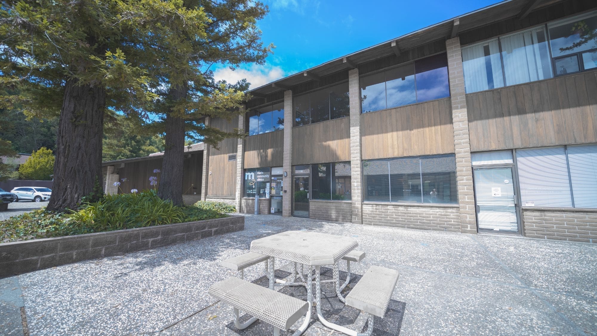 A concrete picnic table with benches sits in front of a two-story office building with large windows, brick pillars, and shaded by tall trees on a sunny day.