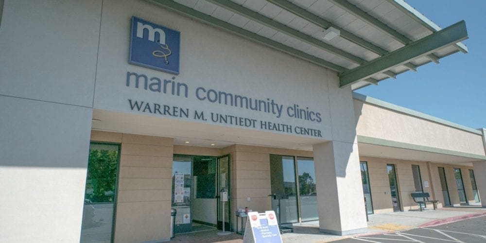 Front entrance of Marin Community Clinics, Warren M. Unietdt Health Center, with glass doors, signage above, and a shaded overhang on a bright sunny day.