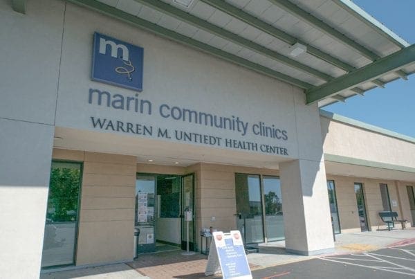 Front entrance of Marin Community Clinics, Warren M. Unietdt Health Center, with glass doors, signage above, and a shaded overhang on a bright sunny day.