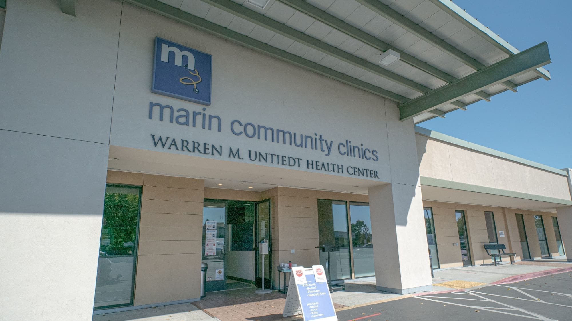 Front entrance of Marin Community Clinics, Warren M. Unietdt Health Center, with glass doors, signage above, and a shaded overhang on a bright sunny day.