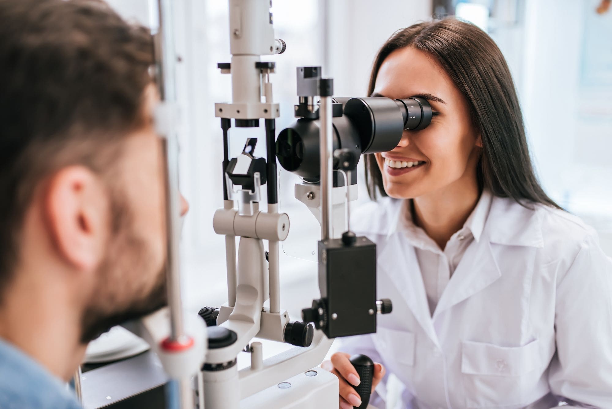 A female optometrist in a white coat examines a male patient’s eyes using a slit lamp in a bright, modern clinic.