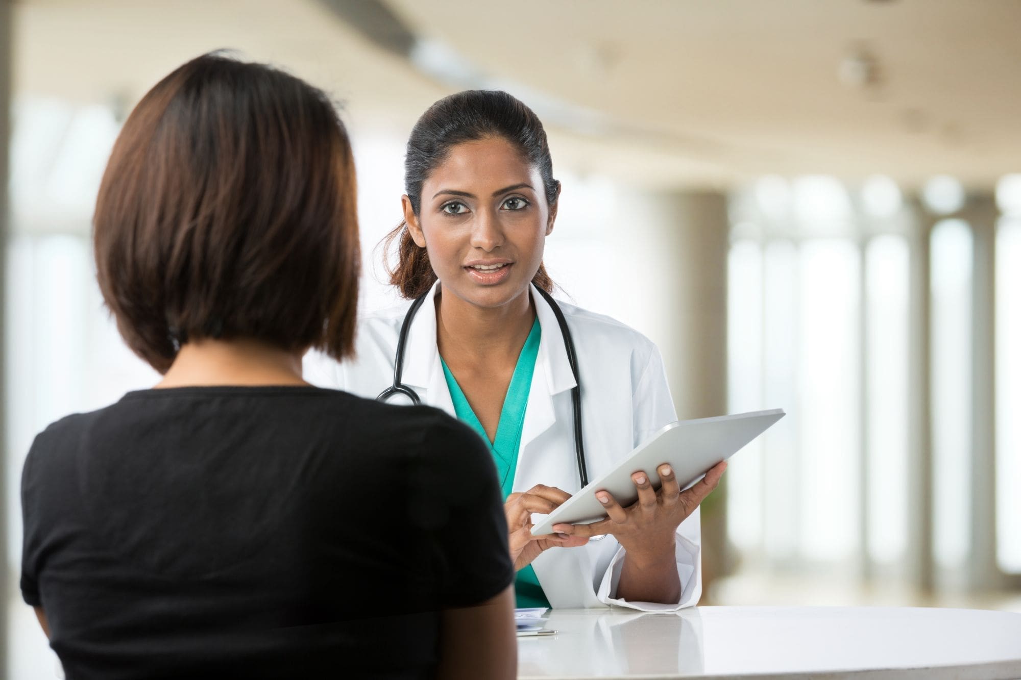 A doctor in a white coat holds a tablet and speaks with a patient whose back is to the camera. They are seated indoors in a bright, modern setting.