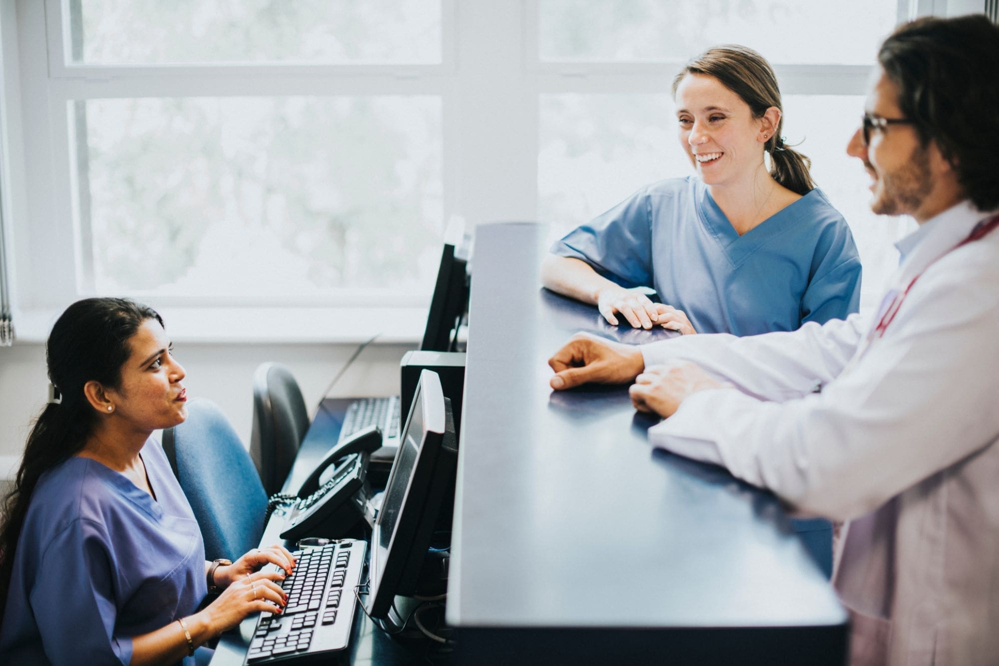 Three medical professionals in scrubs and a lab coat talk at a hospital reception desk; one woman sits typing at a computer while the other two stand and smile on the opposite side of the counter.