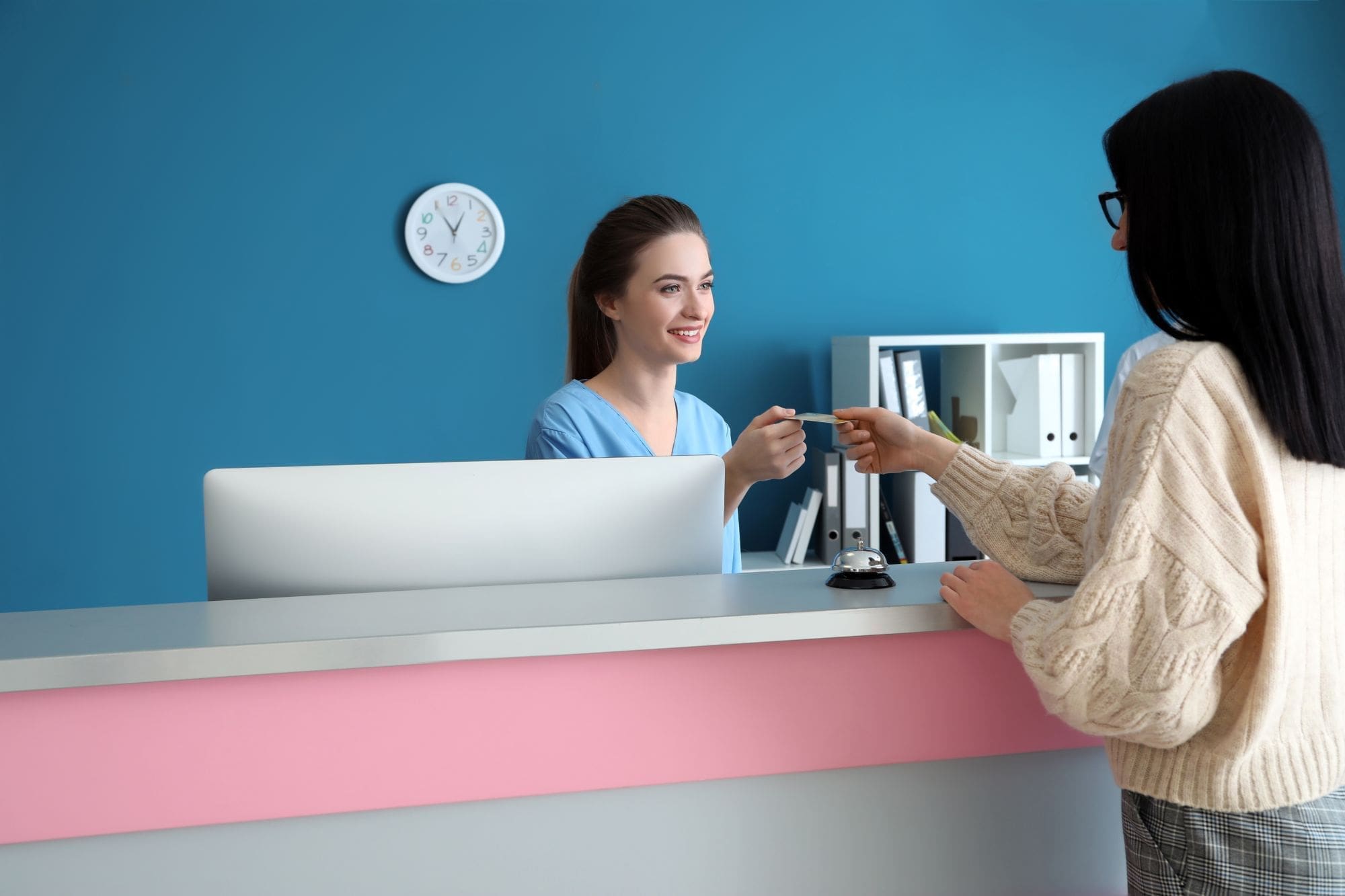 A receptionist in blue scrubs smiles as she hands a card to a woman in a cream sweater standing at a modern front desk with a pink stripe, against a blue wall with a clock and shelves in the background.
