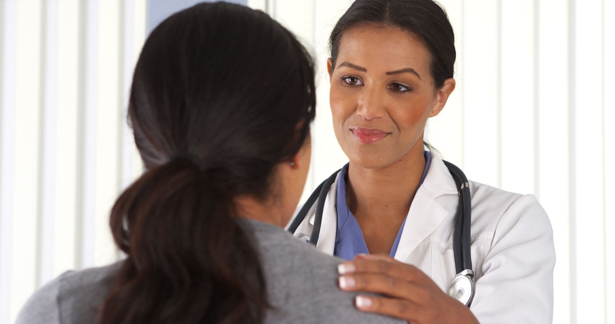A female doctor with a stethoscope gently places her hand on a patient’s shoulder, offering comfort and reassurance. The patient is seen from behind.