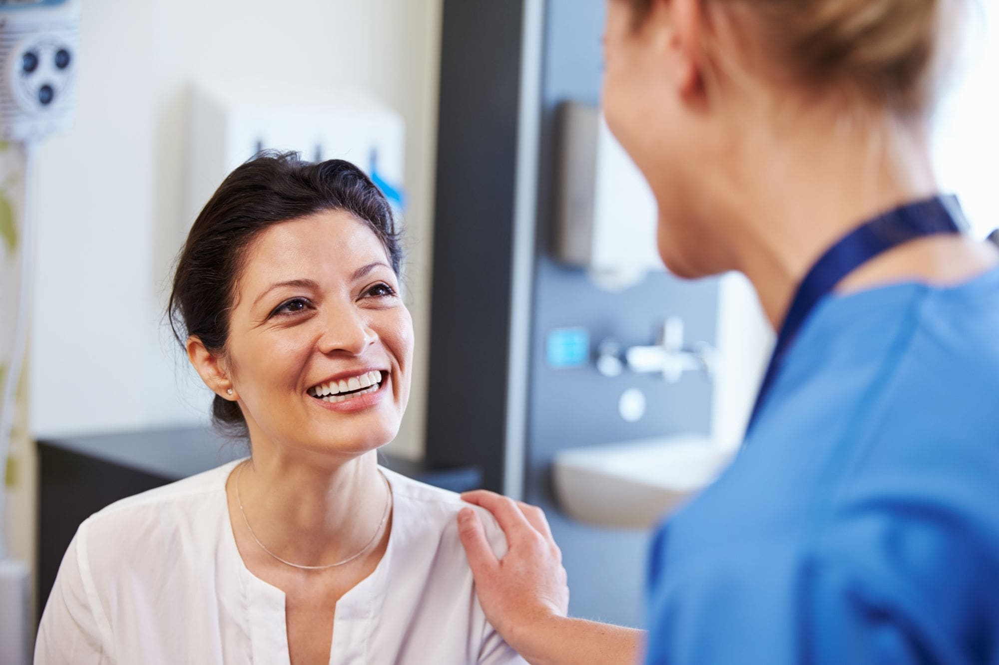 A woman smiles warmly while sitting in a medical office as a healthcare professional in blue scrubs reassures her with a hand on her shoulder.
