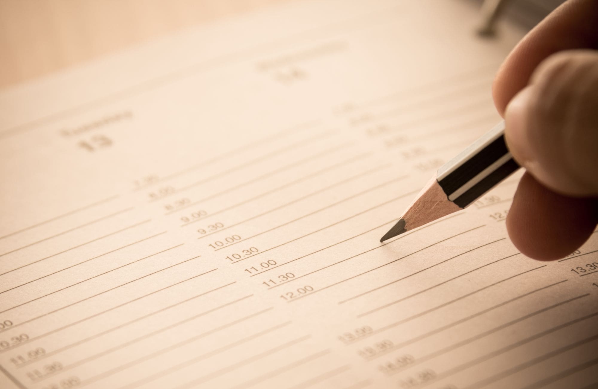 A close-up of a hand holding a pencil above an open planner or appointment book, with time slots listed from 8:00 AM to 12:00 PM, ready to be filled in.