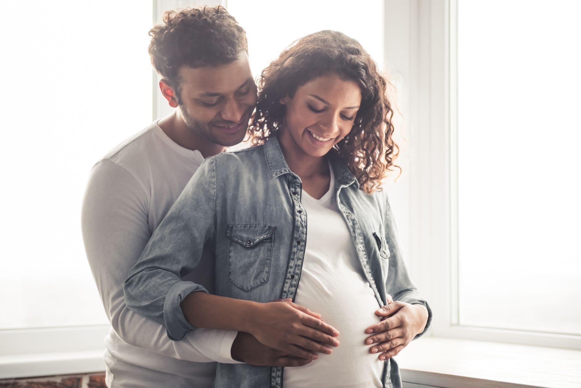 A smiling couple stands by a window. The man embraces the woman from behind, resting his hands on her pregnant belly, while she smiles and gently holds his hands. Both look happy and relaxed.