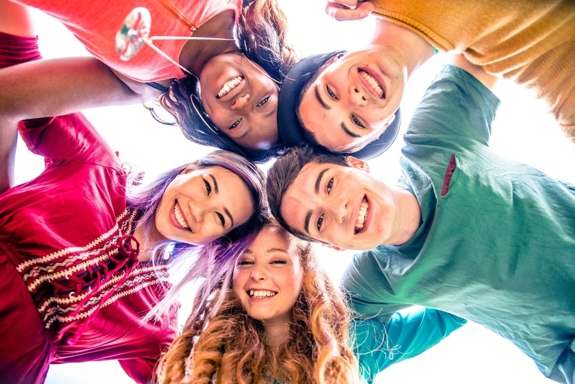 Five young adults stand in a circle, smiling and looking down at the camera. Their arms are around each others shoulders, and they appear happy and cheerful, set against a bright, white background.