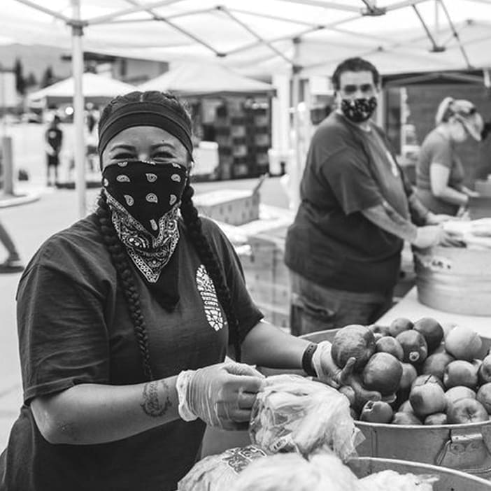 A woman wearing a mask, bandana, and gloves smiles while bagging produce at an outdoor market or food distribution event. Other masked volunteers work in the background under canopies.