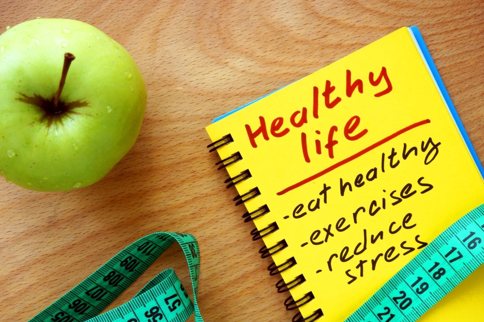 A green apple, a blue measuring tape, and a yellow notepad with Healthy life written on it, listing tips: eat healthy, exercise, and reduce stress, all placed on a wooden surface.